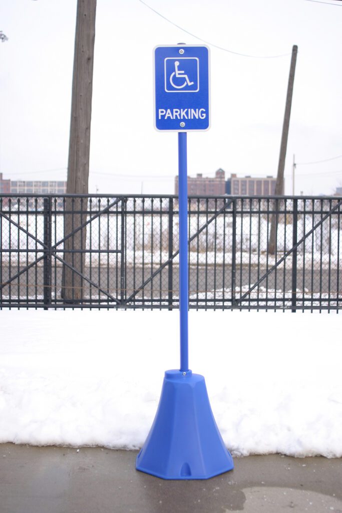 Against a snowy backdrop with a fence, the tall parking sign featuring a wheelchair symbol for accessible parking stands securely on the Vestil Octagon Sign Base Blue 8 X 8 X 98.