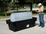 A person in a white hard hat and tan shirt holds a clipboard next to the Vestil Fuel Tank Containment W/ Drain 275 Lb, placed inside a black tray. Trees line the road in the background, completing this industrial scene.