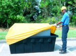 A person wearing a hard hat and gloves adjusts a Vestil Containment Sump Otc-275 Pull Over Cover on a large black container outdoors, with trees and grass enhancing the natural backdrop.
