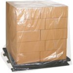 A stack of brown cardboard boxes sits on a black pallet, secured with MH-USA 48 x 42 x 48 - 4 Mil Clear Pallet Covers (25/Case) for added durability.