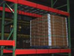 A wooden pallet loaded with boxes is secured by Vestil Nylon Pallet Rack Netting 111 X 48 In on a green and orange warehouse rack. Additional pallets are seen on shelves in the dim background.