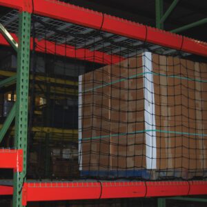 A wooden pallet loaded with boxes is secured by Vestil Nylon Pallet Rack Netting 111 X 48 In on a green and orange warehouse rack. Additional pallets are seen on shelves in the dim background.