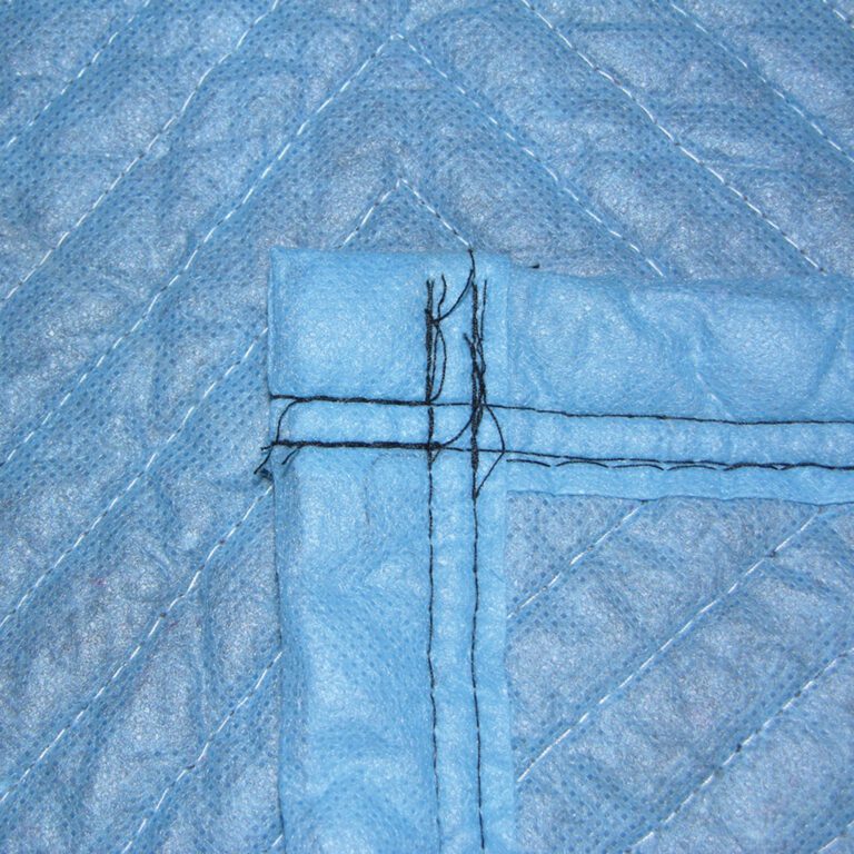 Close-up of a quilted blue fabric corner, similar to Vestil General Duty Moving Pads Cotton 1 Pack. Black stitches intersect at right angles, with textured cotton material and diagonal seams creating a braided pattern.