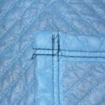 Close-up view of a blue quilted fabric with diagonal stitching and a corner seam, resembling the durable texture of Vestil General Duty Moving Pads Cotton 4 Pack. Intersecting lines and black threads enhance its moving blanket appearance.