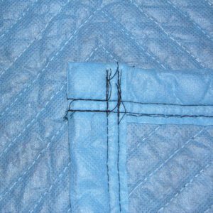 Close-up view of a blue quilted fabric with diagonal stitching and a corner seam, resembling the durable texture of Vestil General Duty Moving Pads Cotton 4 Pack. Intersecting lines and black threads enhance its moving blanket appearance.