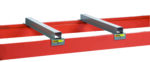 Two gray rectangular metal brackets attach to a red metal beam, resembling parts of the Little Giant Rack Deck Clearance Bar. Yellow caution labels enhance safety in this industrial assembly, with a white background highlighting its structured precision and functionality.