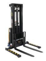 The Vestil Adj Stacker W/Pwr Lift 137In Power Traction Drive is a black industrial pallet stacker with dual long forks, a vertical lifting mechanism, control levers, warning labels, and yellow-black safety stripes on the forks.