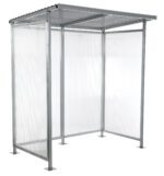The Vestil Smoking Shelter Bus Stop is a compact metal and polycarbonate structure with a slanted roof and three sides. It features a minimalist design, light gray frame, clear panels, and stands on four square bases.