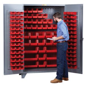 A man confidently stands before a sleek gray Little Giant High Capacity Storage Bin Cabinet, wearing a blue shirt, jeans, and brown shoes. Holding a clipboard, he inspects the neatly arranged multiple red bins inside the cabinet.