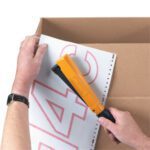 Using a Bostitch Stapling Hammer, someone skillfully tacks perforated paper with red numbers onto a cardboard box, steadying the paper with their left hand and ensuring precise placement with their right.