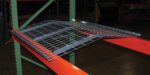 The Vestil Pallet Rack-Crown Wire Deck 52 X 42 In is set on orange beams and a green rack, forming a sturdy platform for items in the dim warehouse.