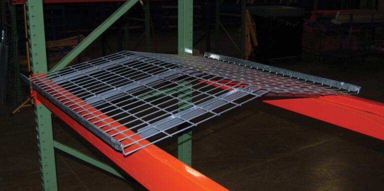The orange and green warehouse pallet rack holds a Vestil Pallet Rack-Crown Wire Deck 58 X 47.6 In. Its slanted wire mesh, detachable design, and versatility are emphasized. The warehouse floor serves as a backdrop.