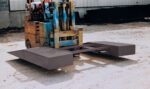 Operating a forklift with a custom attachment, featuring a sturdy metal frame and large flat panels, the operator utilizes the Vestil Wheel Riser Option - Pickup Loop on the dirt surface near the building for better stability during loading.