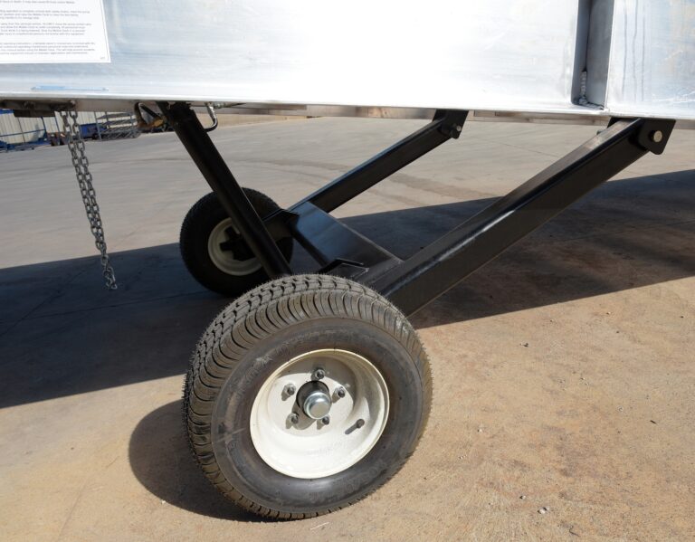 Close-up of Vestil Pneumatic Tire Option on a metal trailer: Two rubber tires with white rims are connected by black metal braces, with a chain hanging from the side, all on a concrete surface.