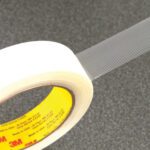 A roll of clear tape with visible fiberglass strands is partially unrolled, showing intricate threads and a yellow label with red text. This is the durable 3M™ 862 Strapping Tape, available in a 1/2 x 60 yds. (12 Pack) per case, set against a dark gray background.