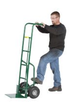 Wearing safety glasses, a person in a black sweater and blue jeans expertly operates the Little Giant 60 Tall Hand Truck with Patented Foot Kick, placing one foot on the lower bar and pushing the handles downward.