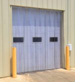A warehouse door showcases the Vestil Vinyl Strip Door 0.12 in full (144 x 120) with clear plastic strips and three small rectangular windows. The beige metal exterior is secured by two yellow bollards, while a wall-mounted mailbox can be seen on the right.