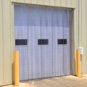 A beige industrial building displays a Vestil Vinyl Strip Door, 0.08 inches thick, measuring 84 x 72 inches, featuring a large transparent curtain with three small rectangular windows, two yellow bollards, and a white box mounted on the right side.
