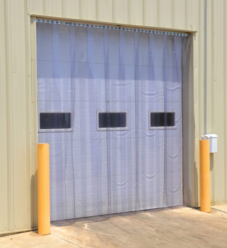 A beige industrial building features a large roll-up garage door with three rectangular windows and a Vestil Standard Vinyl Strip Door 72 X 108. The entrance is protected by two yellow bollards, and light brown pavement surrounds the scene.