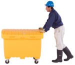 A person in protective gear, including a blue helmet, dark shirt, white pants, gloves, and black boots pushes a large yellow Vestil Utility Box with rubber wheels.