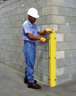 A construction worker in blue jeans, a light blue shirt, gloves, and a white helmet is installing a Vestil Poly Corner Protector on a concrete wall in an industrial or construction area.