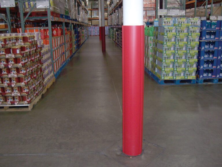 VCW-RD-RND_A.jpg A warehouse aisle with shelves of packaged goods is centered by a Vestil Red Round Column Wrap 8 In, extending from floor to ceiling. The concrete floor stretches to the end of the aisle, enhancing visibility.