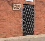 A Vestil Galvanized Scissor Gate-Expanded 67 In spans a brick wall doorway, its diamond patterns adding a metallic sheen in contrast to the red bricks. Above it, a sign reads Office Entrance, while the ground below is neatly paved.
