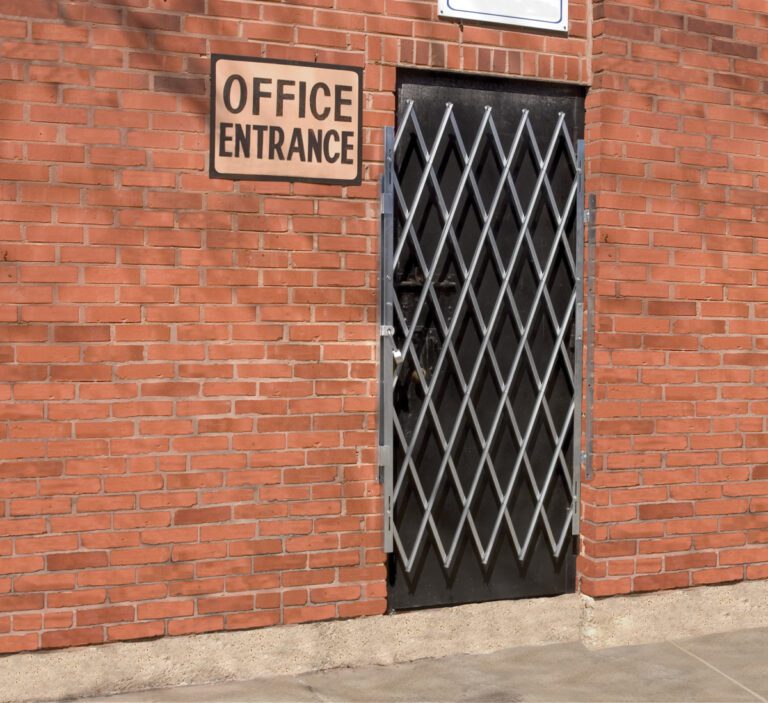 A Vestil Galvanized Scissor Gate-Expanded 67 In spans a brick wall doorway, its diamond patterns adding a metallic sheen in contrast to the red bricks. Above it, a sign reads Office Entrance, while the ground below is neatly paved.