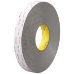 A partially unrolled roll of 1/2 x 5 yds. gray 3M™ 4926 VHB™ tape reveals its adhesive side, with a white cover featuring red 3M logos and a distinctively yellow inner core.