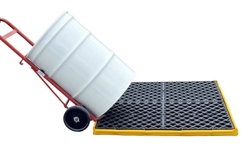 A white barrel rests on a red hand truck, close to a Vestil Ultra Spill Deck Four Drum Flexible. The black and yellow spill platform features a grid pattern to prevent spills from reaching the ground.