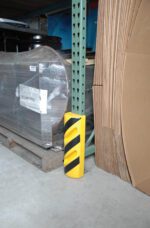 A Vestil Poly Corner Protector 15-1/2 In Height, a yellow and black striped safety guard, wraps around the base of a warehouses metal shelving unit. Nearby lie stacks of wrapped pallets and large sheets of cardboard.