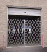 A Vestil Double Fold Scissor Gate Galv 240L X 84H is closed in front of a partially open garage door, surrounded by a beige brick structure with two red bollards on each side.