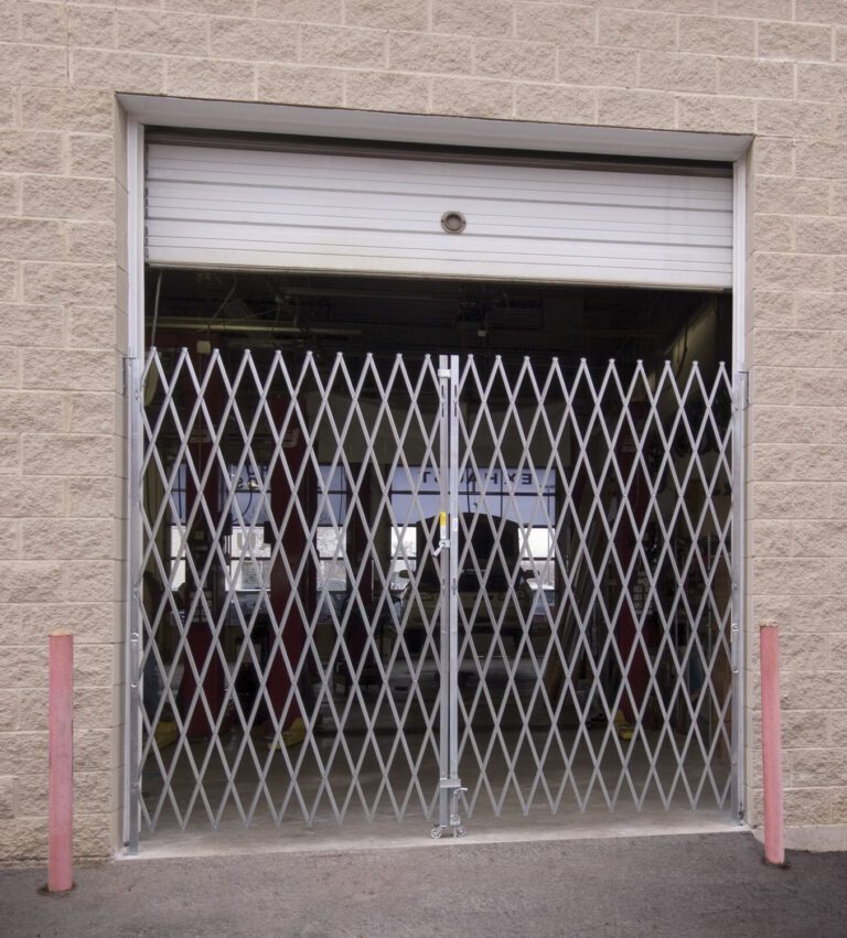 In front of an open garage door set in a light brick wall, a Vestil Double Fold Scissor Gate Galv 96L X 90H is closed. Two red bollards stand beside the sturdy metal gate, revealing part of the interior.