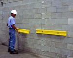 Wearing a hard hat and safety glasses, a person installs a Vestil Polyethylene Wall Protector 48 In on a gray cinder block wall using a tool, while another yellow bumper rail is already secured beside it.