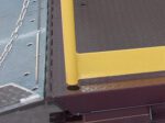 Close-up of a metal platforms corner with a yellow safety edge and diamond plate pattern, featuring the hinge and chain on a smooth surface, enhanced by the Vestil Dock Lift Optional Handrail W/ Toeboard for added safety.