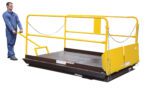 A person in blue overalls operates a lift platform with yellow rails and a handle. The platform, featuring protective chains and safety barriers, includes the Vestil Dock Lift Optional Mobile Wheel Kit for better mobility on its diamond-patterned metal surface.