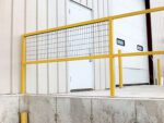 An 84-inch Vestil Steel Sq Safe Handrail Wire Mesh is mounted on a concrete platform outside a beige industrial building. A white door, a large garage door, and yellow bollards are visible in the background, enhancing the secure environment.