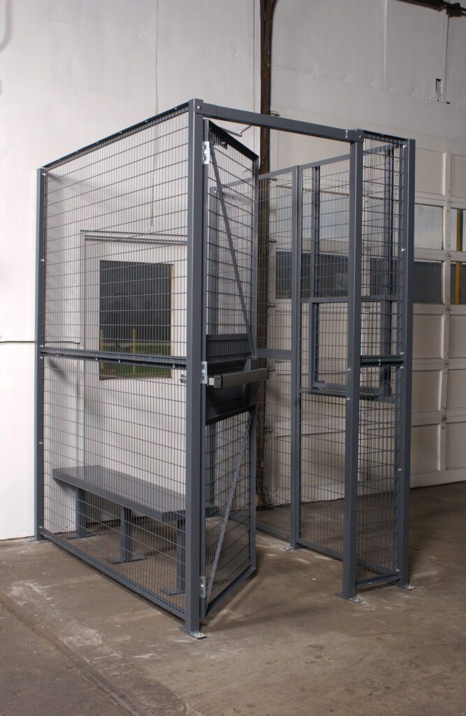 WPC-D-4X4-3_A.jpg A Vestil Driver/Trucker Access Cage 3 Sided 4X4, featuring a sturdy metal door and wire mesh sides enclosing a bench, is tucked in the corner of a room with concrete floors and pristine white walls.