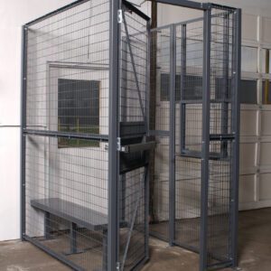 A Vestil Driver/Trucker Access Cage 3 Sided 4X4, featuring a sturdy metal door and wire mesh sides enclosing a bench, is tucked in the corner of a room with concrete floors and pristine white walls.