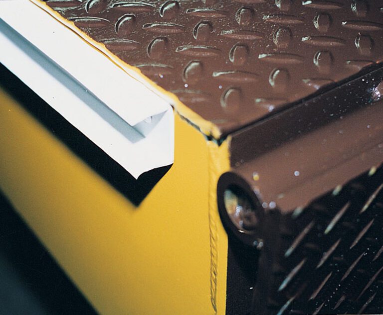 Close-up of a yellow and brown metal surface with a diamond plate texture features white foil resembling the Vestil Optional Dual Vinyl Weather Stripping along the edge. A cylindrical hinge appears on the right side.
