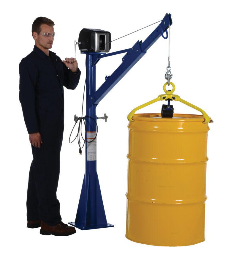 A person in a dark blue jumpsuit operates a Vestil AC Power Lift Jib Crane 1.5K with a 66-inch boom, hoisting a large yellow drum using the hand crank and hook, suspending it mid-air.