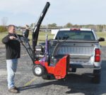 A person uses a hand-cranked lift to load a red snowblower into a pickup truck bed with the Vestil Hitch-Mounted Truck Jib Crane. The truck is in an outdoor lot under a clear blue sky.