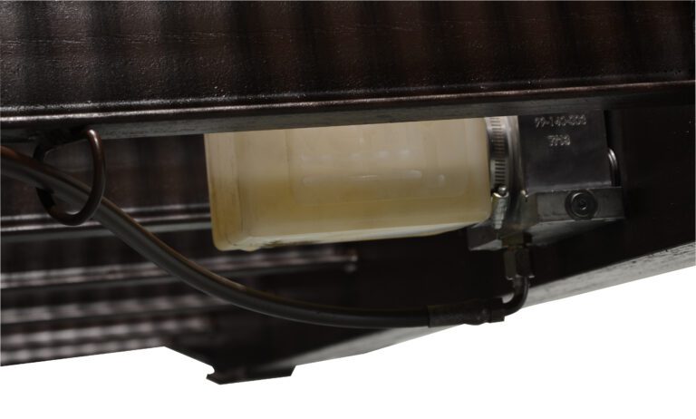 Close-up of Vestil Yard Ramp Option Pnu Tire & Manual Lift: a mechanical component with a translucent plastic container, metallic parts, and black cables on a corrugated surface, depicting an industrial setting.