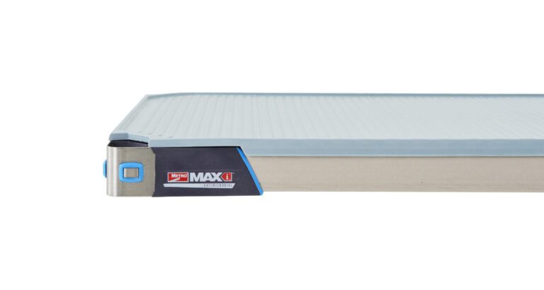 Close-up of a gray and blue industrial platform trolley with a flat surface and reinforced corners. The side displays a MAXX logo and an LED indicator. The trolley is designed for heavy-duty transport and warehouse use.