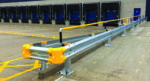 A loading dock area with multiple blue loading doors, surrounded by a safety barrier. The barrier is made of metal with yellow protective bumpers. The ground is marked with yellow lines to indicate parking or maneuvering areas.