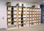 Shelves filled with labeled brown cardboard boxes in an office setting. The boxes are arranged in rows, each labeled with text on the front. The room has a white wall and door, with a carpeted floor.