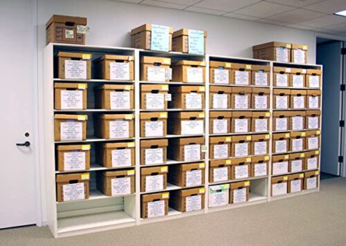 Shelves filled with labeled brown cardboard boxes in an office setting. The boxes are arranged in rows, each labeled with text on the front. The room has a white wall and door, with a carpeted floor.
