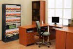 Office with wooden furniture, including an L-shaped desk, swivel chair, filing cabinet, and bookshelf filled with files. A computer monitor is on the desk, and a large window provides natural light from the right.