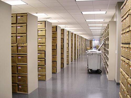 An archive room with bright lighting features Aurora Quik-Lok Shelving, adjustable heavy-duty steel units with 350 lbs capacity. Stacked brown boxes line the shelves, enhancing a clean atmosphere on a gray floor and white ceiling, while a large gray cart stands at the center.