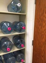 Five large water bottles with pink caps are stored horizontally on the sturdy beige Aurora Quik-Lok Shelving unit, which boasts adjustable shelves and can hold up to 350 lbs. A wooden door is slightly open on the right side, revealing a white painted wall behind it.